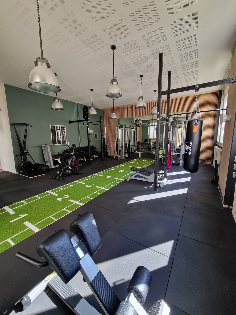 Vue d'ensemble du studio Top Strength à Lyon 6 : piste de sled en gazon synthétique, rack, haltères, sac de frappe et mur en lattes de bois